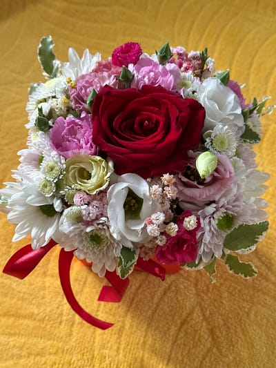 A close-up, overhead shot displays a vibrant bouquet of mixed flowers arranged in an orange box, tied with a red ribbon, resting on a textured yellow surface. The bouquet features a prominent, deep red rose at its center, surrounded by various pink and white flowers, including carnations, chrysanthemums, and smaller delicate blossoms. Green leaves are interspersed throughout. The yellow surface appears to be a soft fabric with a subtle pattern. The image does not contain any elements that would indicate its location in the city of Wisła, Poland.