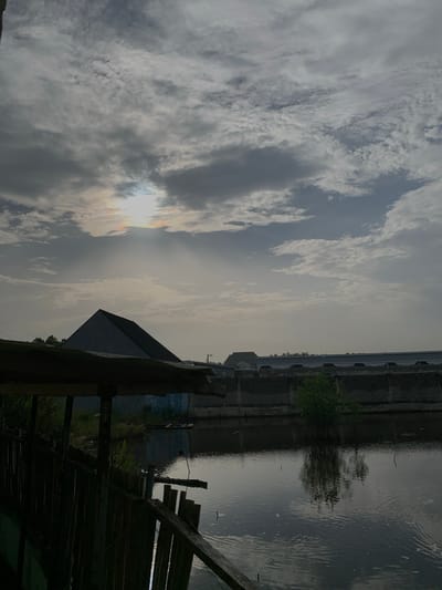 The image captures an overcast waterside landscape in Ibasa, Nigeria. The upper two-thirds of the frame are filled with a dramatic sky, featuring extensive grey and white clouds. The sun is visible as a bright, diffused light source breaking through the clouds, creating a luminous spot with subtle iridescent hues around its edges.

Below the expansive sky, a calm body of water, likely a river or canal, occupies the lower third of the image. The water reflects the subdued light of the sky, showing gentle ripples on its surface. On the far bank of the waterway, a small, bushy green tree stands, while a long, low concrete wall or embankment lines the water's edge further along.

To the right, a distinctive, dark pyramidal building with a sharp, angular roof is prominent, its structure silhouetted against the bright sky. Other distant buildings and structures are faintly visible in the background, suggesting a developed area along the water. In the immediate foreground on the right, dark wooden structures, appearing to be parts of a jetty or platform, extend into the scene.

The lighting suggests an overcast day, possibly late morning or afternoon, with the sun's light diffused by the cloud cover. The scene is devoid of human activity, conveying a quiet and serene atmosphere. There is no visible text in the image.