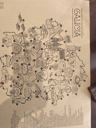 The image displays a detailed, hand-drawn map of the autonomous community of Galicia, Spain, presented on a textured, light beige surface resembling paper or cardboard. The title "GALICIA" is prominently displayed in black capital letters on the top right.
The map is a compilation of numerous numbered black and white line illustrations representing significant cultural, natural, and gastronomic features of the region. Subjects depicted include architectural landmarks like the Cathedral of Santiago de Compostela (14), the Tower of Hercules in A Coruña (23), and traditional Galician structures such as Hórreos (19) and Pallozas (32). Natural attractions, like the Fragas do Eume (16), Os Cañóns do Sil (34), and Playa de As Catedrais (31), are also illustrated.
A substantial portion of the map is dedicated to Galician cuisine and produce, showing items like Pimientos de Padrón (20), Pulpo de Carballiño (38), Queso de San Simón (25), Tarta de Santiago (18), and a bottle labeled "ALBA RIÑO" (10) representing Albariño wine. Illustrations also highlight local festivals and traditions, including the Fiesta Vikinga (1) and A Rapa das Bestas (4).
Each illustration is correlated with a numbered legend listed on the right and bottom right of the image, providing specific names in Spanish for all 52 depicted items, from "1) Fiesta Vikinga, Catoira" to "52) Magosto, Oure". A "100% RECYCLABLE" logo is visible in the top left corner. The overall style is whimsical and informative, serving as a charming visual guide to the diverse attractions of Galicia.