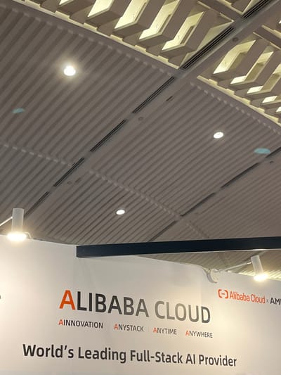 A skewed, close-up, indoor shot displays a sign for Alibaba Cloud and a textured ceiling. The sign, located on the right, is white and features black lettering except for the word "ALIBABA" which is in orange. The sign reads "Alibaba Cloud × AM," "ALIBABA CLOUD," "AINNOVATION ANYSTACK | ANYTIME ANYWHERE," and "World's Leading Full-Stack Al Provider." The ceiling is composed of rows of textured panels with recessed lighting fixtures. The location in the city of Hong Kong, China, is not ascertainable from the image.