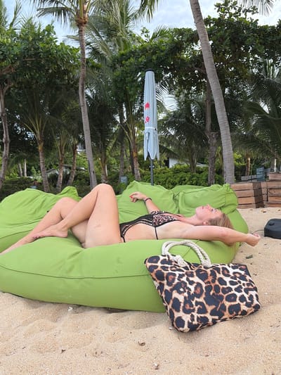 The image shows a woman lying on a large, bright green beanbag on a sandy beach. She is wearing a black and leopard print bikini and has her arms outstretched above her head. Her eyes are closed and she appears to be relaxing. To her right, a leopard print beach bag with rope handles rests on the sand. In the background, there are palm trees, lush green foliage, and a light-colored umbrella. Wooden crates are stacked behind the beanbag. The overall setting suggests a tropical resort location.