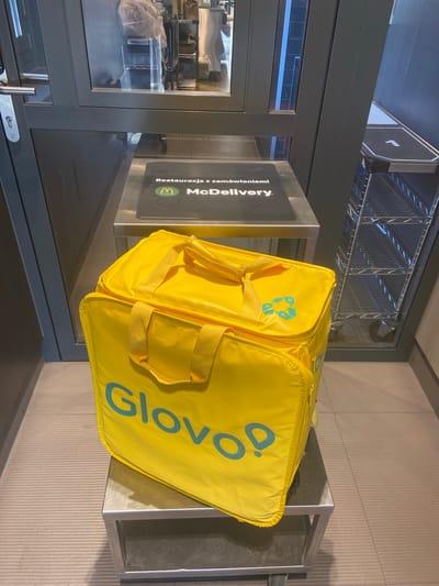 A yellow, insulated delivery bag with the Glovo logo is placed on a metallic shelf. The bag has two yellow handles and a small, turquoise recycling symbol on its side. Next to it is a black sign with a white border, which reads "Restauracja z zamówieniami McDonald's" and features the McDonald's logo. The background consists of a metal shelving unit and a wall with vertical textured panels. The scene appears to be indoors, possibly in a McDonald's restaurant in Bolesławiec, Poland, where food deliveries are processed. There are no people visible in the image.