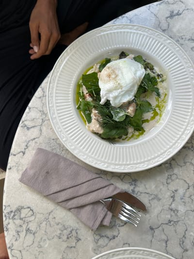 An overhead shot shows a white, ribbed plate filled with a dish of greens, sauce, and a poached egg. A person's hand, with dark skin and light-colored fingernails, rests on black fabric to the left of the plate. A folded gray napkin and a fork and knife set are placed to the bottom left of the plate on a marble-patterned surface.