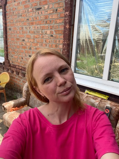 In an outdoor setting, a woman with blonde hair and fair skin is captured in a selfie. She is wearing a bright pink t-shirt. The background features a brick wall on the left and a window with reflective sheeting covering it on the right. A patterned armchair is visible behind the woman, with a yellow chair to the left of it. The overall scene appears to be in a residential area.