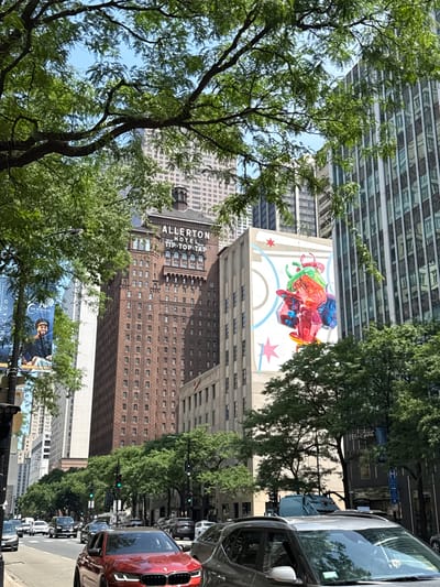 The image depicts a street scene in Chicago, USA, on a sunny day. A line of cars is parked along the right side of the street, including a prominent red car in the foreground. Trees with lush green foliage line the street, obscuring parts of the surrounding buildings.
Several buildings are visible, including a tall, modern building with a glass facade and a brick building labeled "ALLERTON HOTEL TIP-TOP-TAP". A mural of colorful, translucent geometric shapes is painted on the side of one building. Below this, a banner features a man's portrait and the text "THE PENINSULA" and "CHICAGO". The sky is blue and clear. The scene appears to be in a bustling urban environment.