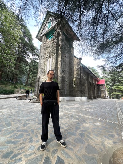 A man stands on a stone pathway in Dharamkot, India. He is oriented horizontally and is wearing a black t-shirt, black pants, and black and white sneakers, with a black fanny pack around his waist. His hair is dark and pulled back. Behind him is a stone church with a red roof and various windows. The church is flanked by trees and foliage. The sky is visible through the trees. In the background, other people can be seen walking along a path. The stone pathway is decorated with patterned tile.