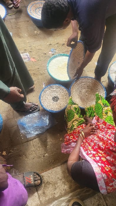 The scene depicts an informal market or food preparation area in Fagge D1, Nigeria. A standing individual, dressed in a dark shirt and trousers, is leaning over, holding a circular woven lid with blue trim over a light-colored granular substance contained within a larger, shallow, round woven basket with blue trim. To the left, a person wearing green attire and sandals is partially visible. On the ground in the lower-right quadrant, a person is seated, wearing a red and yellow patterned dress over dark leggings, with gold bracelets on their right wrist. Two other similar woven baskets, one with a prominent concentric pattern, are positioned near the seated individual. A clear plastic bag containing an indistinct dark object rests on the dirty, uneven concrete ground. A foot wearing a black "ZARA" slide sandal is visible in the bottom left corner. The environment is characterized by a dusty, somewhat cluttered ground surface.