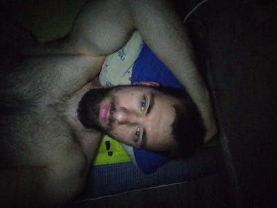 An overhead, low-angle, night-time, close-up shot shows a man lying on his back with his head resting on a pillow and a blue blanket. The man has dark hair and a beard, and his eyes are open and looking at the camera. He is wearing a white sock on his left foot, which is partially visible. The pillow has a yellow pattern with a black exclamation point. The man's left arm is visible, and he appears to be shirtless. The lighting in the scene is dim, casting shadows and obscuring some details. The overall impression is of a casual, indoor setting, possibly a bedroom. There is no information in the image to indicate the location in Gyeongju-si, South Korea.