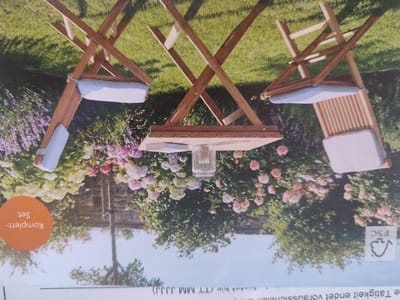The image displays a wooden folding table with a glass vase or container on it. Two wooden folding chairs with white cushions are positioned on either side of the table. The scene is set outdoors on a grassy area, with abundant pink and white flowers in the background, suggesting a garden or park setting. The overall impression is one of a relaxed, outdoor seating arrangement. There are no people present in the image. The image is rotated such that the text is upside down. The text in the orange circle on the left reads "Komplett-Set". The text at the bottom right reads "FSC".