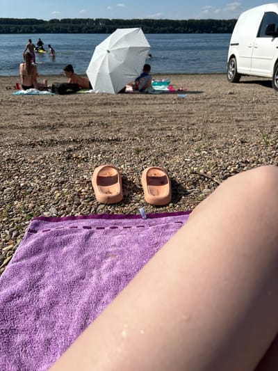 The image captures a sunny day at a pebble beach along the Danube River in Lom, Bulgaria. In the foreground, a purple towel lies on the pebbled shore, with a pair of light brown slip-on shoes resting on the pebbles nearby. A person's leg, with visible water droplets, occupies the lower right portion of the frame, suggesting they are either sunbathing or have recently been in the water.
In the mid-ground, several people are visible on the beach. Two women are seated on towels, one in a black bikini and the other in a pink bikini, with a beach bag between them. A white beach umbrella provides shade. Further back, a group of people are in the water. One person is on a jet ski, while others appear to be swimming or wading.
The background features the calm waters of the Danube River, with a dense line of trees along the opposite bank. A white van is parked on the beach to the right of the frame, its tires visible on the pebbles. The sky is blue with scattered white clouds. The overall atmosphere is one of leisure and recreation.