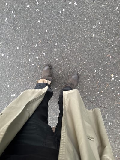 A person stands on an asphalt surface scattered with small white spots, possibly flower petals, indicative of springtime. The individual wears dark trousers and brown leather boots. A light-colored trench coat, buttoned at the top, is draped around them. The location is likely an outdoor area in Les Ulis, France, based on image context.
