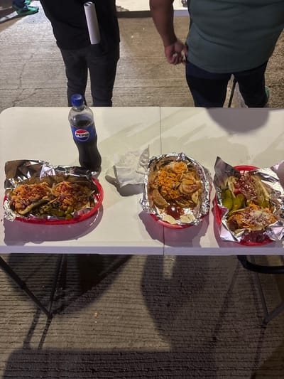 The image shows a table with food and drinks, likely in Tampico, Mexico. Three red baskets contain food wrapped in aluminum foil; two appear to be sandwiches or tacos with toppings like shredded cheese, diced tomatoes, and pickles, while the middle basket holds what looks like nachos covered in shredded cheese or a similar topping. A bottle of Pepsi is visible on the table next to one of the food baskets, along with a white napkin. Parts of two individuals are visible around the table - one wearing a green shirt and the other wearing black pants. The table is on a concrete surface.