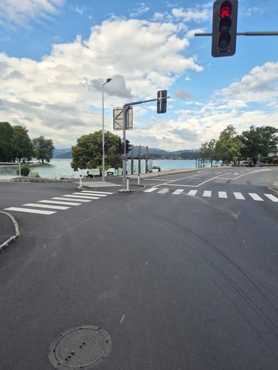 The image captures a view of an intersection at a body of water, likely a lake, in Seewalchen am Attersee, Austria. A prominent traffic light with a red arrow pointing left is visible in the upper right portion of the frame, indicating a stop or turn restriction. Across the road, a pedestrian crossing with white stripes is marked. The road surface is asphalt, with some tire marks visible. In the background, trees line the shore, and the water stretches out to a distant, hazy shoreline with hills. The sky is a mix of blue and white clouds. A signpost reads "POLIZEI," indicating a police presence. The overall scene suggests a quiet, possibly resort town setting.
