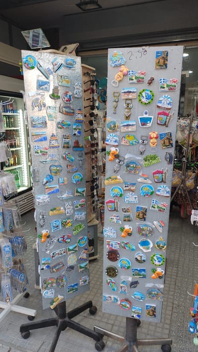 The image shows two tall, grey, rotating display stands filled with numerous colorful souvenir magnets. The stands are positioned outdoors, with a tiled ground surface visible and some shelving units with items on the left. In the background, a building facade and a partial view of a shop interior are present. The magnets display various scenes and objects, many of which appear to be related to Greece, including lighthouses, windmills, ships, beaches, and traditional Greek motifs. Some magnets also feature text, with "Greece" and place names like "Chalkidiki" and "Thessaloniki" being discernible. The display stands are on wheeled bases, suggesting they are mobile. There are no people visible in the image. The setting appears to be a souvenir shop or a market stall in Perea, Greece.