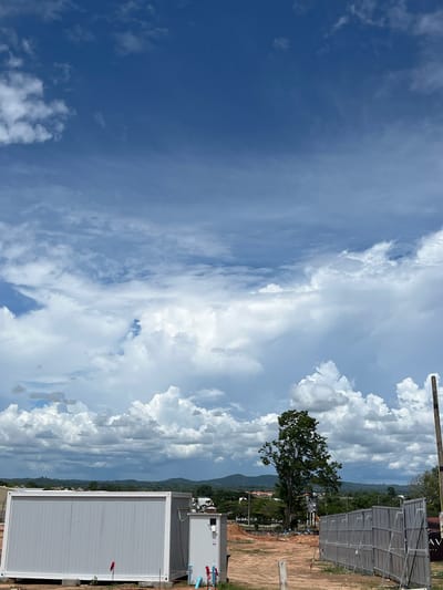 The image shows a landscape in Muang Pattaya, Thailand, under a partly cloudy sky. To the right, there's a temporary structure, possibly a container building, and a partially visible metal fence structure. The land appears to be undergoing construction or renovation. A tree is visible near the fence. Mountains and buildings appear in the background horizon. The sky on the left side has a deep blue hue and scattered clouds, contrasting with the right side's more dense cloud cover.