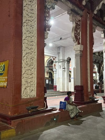The image depicts an interior scene, likely a temple or historical building in Chennai, India, given the architectural style and the presence of a woman in traditional attire. The time appears to be evening or night, as indicated by the artificial lighting and the deep shadows cast on the ground.
The main subject is a woman sitting on a raised platform, leaning against a red pillar. She is wearing a purple blouse and a light-colored sari draped over her. She holds her hands together, appearing contemplative or resting. Beside her is a blue bag. The setting is characterized by ornate architectural details, including carved friezes on the walls, decorative pillars, and arched openings. The walls are painted in muted tones of white and red, with intricate patterns and embellishments.
In the background, other people can be seen walking in a corridor. The overall atmosphere is serene and somewhat quiet, with the focus on the woman and the architectural grandeur of the location. A sign with text is partially visible at the bottom of the frame. The visible text on the yellow sign appears to be in English and possibly another script, with words like "PROTECTION FORCE" and "PARKING" discernible. The lighting is a mix of bright spotlights and softer ambient light, creating a dramatic interplay of light and shadow.