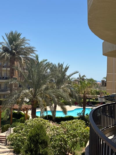 This image captures a sunny day with a clear blue sky, suggesting it's likely daytime and the weather is fair. The scene appears to be a resort or hotel setting, with a swimming pool as a central feature. Lush green trees and palm trees are abundant, indicating a warm climate, possibly Egypt given the location context. A dark metal railing curves along the right side of the frame, hinting at a balcony or elevated walkway. In the background, a building with a beige facade and visible windows suggests accommodation. The overall atmosphere is one of leisure and relaxation, common in vacation destinations. There is no visible text in the image.