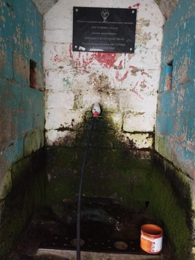 A low-angle, slightly high-contrast medium shot shows the interior of a stone structure, likely a well or a shrine, in Darvag, Russia. The walls are constructed of rough, painted cinder blocks, with areas of peeling teal and white paint, revealing underlying brickwork and stains. Red graffiti, resembling arrows and abstract shapes, is visible on the white walls.
A black plaque with text in Russian is mounted high on the white wall. The text appears to be an inscription related to a cultural heritage site. Below the plaque, a white faucet with a red band is attached to a pipe. A black hose extends downwards from the faucet.
The floor of the structure is dark, wet, and covered in green moss and dark stains, suggesting consistent moisture and neglect. There are some circular indentations or drains visible in the floor.
In the lower right corner, an orange plastic container with white labels, possibly a bucket or can, rests on the floor. The lighting is dim, with some areas appearing brighter due to reflection. The overall impression is one of disrepair and abandonment in a historical or natural setting.