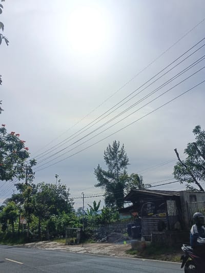 The image depicts a bright, sunny daytime scene along a rural or semi-rural road, consistent with a setting like Carcar, Philippines. The sky is intensely bright, almost appearing white, with the sun prominently visible as a brilliant, washed-out disk in the upper left quadrant. Multiple dark power lines stretch diagonally across the upper part of the frame, originating from a utility pole on the left.
The foreground shows a section of an asphalt road, with a white line visible on its left edge. Both sides of the road are densely covered with green vegetation, including various trees and bushes. A tree on the far left, near the utility pole, features distinctive clusters of orange-red flowers. In the mid-ground, a tall, slender pine-like tree stands out against the lighter sky and other foliage.
On the right side of the road, there is a rustic, single-story roadside stand or small building. It has a corrugated metal roof and appears to be constructed from simple materials like wood or concrete blocks. Several dark barrels or containers are placed outside the structure. A prominent sign on the building clearly displays the text "MAGNUM COFFEE," with another smaller sign below it showing "20.000," likely indicating a price. To the far right, partially visible, a person wearing a helmet is seated on a black motorcycle, suggesting a brief stop or a moment of rest. The scene conveys a sense of a typical, quiet day in a lush, tropical environment.