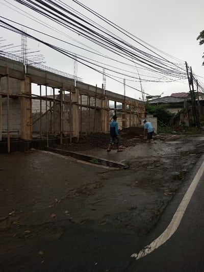 The image depicts an outdoor scene in the city of YonPhoogla, Bhutan, featuring a construction site alongside a wet asphalt road under an overcast sky.
In the foreground, a dark asphalt road, damp and containing scattered debris, runs from the lower left to the right, with a solid white line marking its right edge. A metal drain grate is visible on the left side of the road.
To the left, a single-story structure is under construction. It consists of light-colored block walls, concrete columns, and a concrete beam running horizontally across the top. Wooden scaffolding and temporary supports are integrated into the structure. Exposed rebar protrudes vertically from the tops of several columns. Piles of dark aggregate and loose soil are present on the unpaved ground directly in front of the construction.
Two adult males are visible near the construction. The individual on the left wears a blue long-sleeved shirt and patterned shorts, standing barefoot with his back partially turned. The individual on the right wears a light blue t-shirt and dark shorts, also barefoot, bending over near a small stack of light-colored bricks or blocks, appearing to be engaged in work.
Numerous black utility lines traverse the upper portion of the frame, strung overhead and connecting to a utility pole on the far right. In the background, behind the construction site, residential buildings with tiled roofs are visible amidst green foliage.