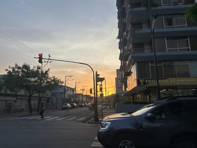 The image shows a street scene in Guayaquil, Ecuador, at dusk. The sky is illuminated with a warm, golden sunset. A pedestrian is crossing a crosswalk on the left side of the frame, while a dark-colored SUV is prominently positioned on the right side of the image. Both traffic lights visible in the scene are displaying red signals. On the right, a building with balconies and a storefront with a sign that includes the words "TROFEOS ECUADOR" stands adjacent to the street. A row of parked cars lines the street in the background, with other buildings and utility poles visible further down the street.