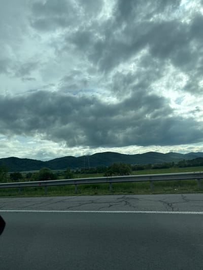 The image depicts a view from a moving vehicle on a road, likely a highway, with a gray asphalt surface and a white dashed line. A metal guardrail separates the road from a grassy embankment. Beyond the embankment, rolling green hills are visible, covered in dense vegetation. The sky is overcast with heavy, dark gray clouds, suggesting an impending storm or a generally gloomy day. In the distance, on the hills, are power lines and what appear to be transmission towers. The overall impression is one of a rural landscape under a dramatic sky. The location is identified as Mirovo, Bulgaria.