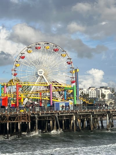 A Ferris wheel with colorful gondolas sits on a pier beside the ocean. The sky is overcast with gray clouds. Buildings line the shoreline in the distance. Waves crash against the wooden pier pilings. Other amusement park rides are partially visible.