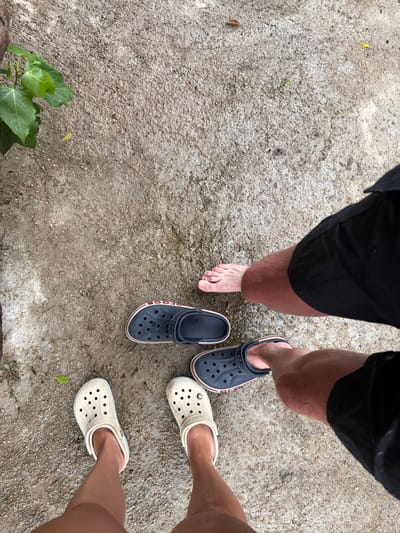 The image shows a top-down view of two pairs of Crocs shoes on a wet, gritty surface, possibly concrete or asphalt, with scattered small stones. In the lower left, a pair of light beige Crocs are on a person's feet. To the right and slightly higher, a pair of dark navy Crocs with red trim are on another person's feet, with one bare foot visible between them. Further up and to the right, another bare foot is visible next to a dark navy Croc. Black shorts are visible on at least two individuals. In the upper left corner, a plant with large green leaves is partially visible. The scene appears to be outdoors, possibly in a public area or backyard in Khanh Vinh, Vietnam, given the texture of the ground and the presence of tropical foliage.
