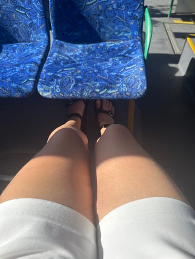 The image depicts the interior of a public transportation vehicle, likely a bus or train, with a focus on the seating area. Two blue patterned seats are visible in the upper portion of the frame, with their distinctive paisley-like design. The lower portion of the image shows a person's legs and feet, with one leg extended forward and the other bent. The person is wearing white shorts and black sandals. Sunlight casts shadows across the legs, indicating the presence of a window. The environment suggests an urban setting, but there is no specific information about the location being Höhenkirchen-Siegertsbrunn, Germany. The overall scene is candid and informal, capturing a moment during a journey.