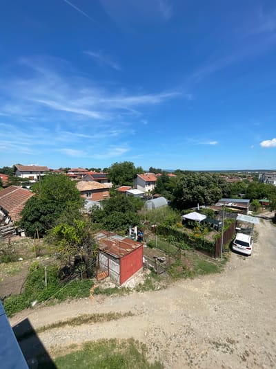 An eye-level, medium shot captures a rural scene on a clear, sunny day in Debelt, Bulgaria. The foreground is dominated by a dirt road, with patches of green grass and scattered stones. To the left, a portion of a blue structure extends into the frame, casting a shadow across the foreground.

The midground reveals a collection of structures and greenery. A weathered, red metal shed with a rusted roof stands prominently in the center, adjacent to a smaller, similarly rusted structure. A thin wire stretches across the scene from left to right, supported by posts. Lush green vegetation and trees fill much of this area, with some appearing to be fruit trees. A greenhouse, covered with translucent material, is visible among the plants.

Further back, the scene transitions to a residential area with houses featuring red-tiled roofs. These buildings are interspersed with more trees, creating a semi-rural, semi-suburban atmosphere. A white car is parked on the dirt road, facing away from the viewer, to the right of the greenhouse. In the distance, more buildings and trees extend towards the horizon. The sky is a vibrant blue, dotted with thin, wispy white clouds and a single, faint contrail. The overall impression is one of a well-lit, peaceful day in a small Bulgarian town or village.