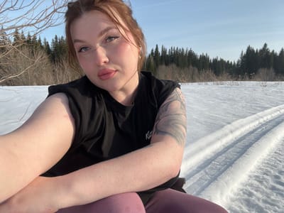 An adult female is depicted in a close-up, self-portrait photograph taken in a snowy, open landscape, likely in the vicinity of Pavlovka, Russia. The subject, with fair skin, reddish-brown hair pulled up, and light-colored eyes, faces the camera directly. She wears prominent eyeliner and light pink lipstick. Her attire consists of a black, sleeveless top and pink or mauve-colored pants. A large, dark-inked tattoo, featuring a swirling or serpentine design, is visible on her left upper arm. The background reveals an expanse of white snow under a clear, pale blue sky. Parallel tracks, consistent with skis or a sled, are imprinted in the snow to the right of the subject. Bare deciduous trees and bushes populate the mid-ground, transitioning to a dense line of dark evergreen trees in the distant background.