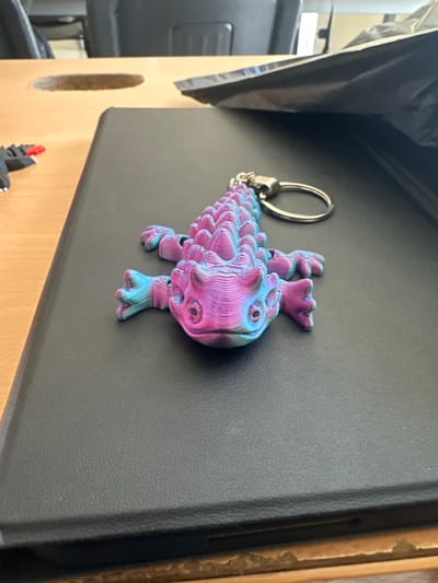 A 3D-printed lizard-shaped keychain, predominantly magenta and light blue with visible print layer lines, rests on a black textured surface, likely a tablet case, in an indoor setting. The lizard keychain features a segmented, spiky back, small circular eyes, and four limbs, with a silver-colored metal keyring attached to its head. The black case is situated on a light-brown wooden table that exhibits a circular cutout on its left side. In the background, dark-colored office chairs are visible, along with a crumpled black plastic object on the table to the right. The scene is located in Fraccionamiento Los Heroes Queretaro, Mexico.