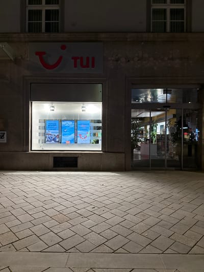 The image shows the exterior of a TUI travel agency in Innsbruck, Austria, at night. The facade is made of stone, with a large window display featuring travel advertisements. One poster is for "Vorfreude garantiert. Sommerurlaub buchen." (Anticipation guaranteed. Book your summer vacation.), another for "WOW ABENTEUER URLAUB" (WOW Adventure Vacation), and a third for "LAST MINUTE SALE." The TUI logo, a red smiley face and the word "TUI," is visible above the window. To the right, a glass door leads into the brightly lit interior of the agency, where plants and a counter are visible. The pavement in front of the building is paved with rectangular bricks.