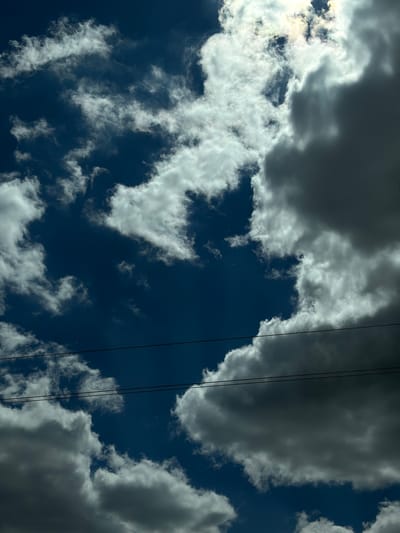 The image depicts a view of the sky above Znamenka, Russia. The sky is a deep blue, partially obscured by a mix of white and grey clouds. The clouds are of various sizes and densities, creating a dynamic pattern against the blue backdrop. Two black power lines diagonally bisect the upper right section of the image.