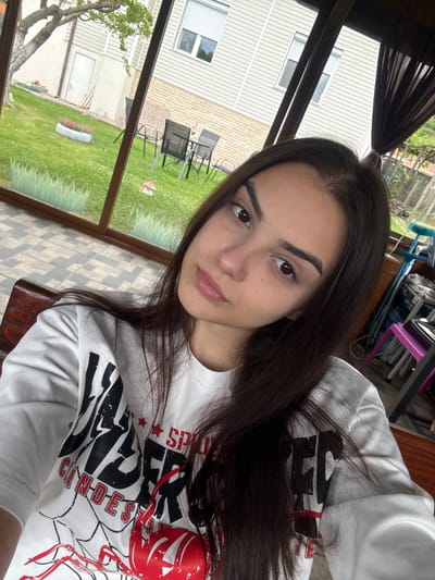 A young woman is taking a selfie indoors. She has dark hair and is wearing a white t-shirt with a graphic design. The background shows an outdoor setting with a house, grass, chairs, and trees visible through a window or screen. The location might be a sunroom or enclosed porch. There's also some furniture and objects in the background behind her, including a blue object, a brown surface, and a pink chair. I am unable to determine the location is the city of Polena, Bulgaria.