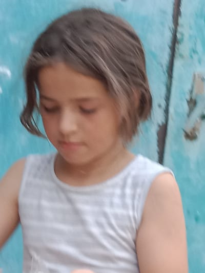A young girl with brown hair is looking down and to her left. She is wearing a white tank top with thin grey horizontal stripes. She has a neutral expression. The background is a textured, weathered blue wall. The lighting suggests it is daytime.