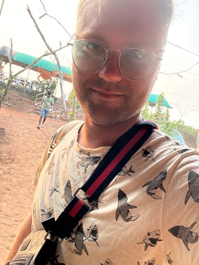 An outdoor, eye-level selfie captures a light-skinned male in the foreground, slightly smiling, wearing round metal-rimmed eyeglasses that reflect the bright sky. He wears a light-colored t-shirt patterned with grey sharks and a cross-body bag with a dark blue and red striped strap. The background features a reddish-brown dirt path under a bright, hazy sky in Bhagatwadi, India. In the middle ground, a second male, with darker skin, wearing a light t-shirt and blue shorts, walks from left to right on the path. Two dogs, one light brown and one darker, are visible near his feet. To the upper left, a multi-level outdoor establishment with a green tarp roof and wooden railings is visible, featuring a large orange crab-shaped sign and partially visible text "BHAI CRAB". Bare tree branches and wires extend across the frame.