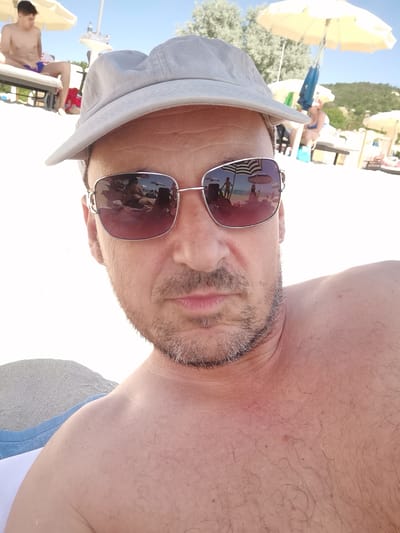 A man in sunglasses and a cap is taking a selfie on a beach. He is wearing a gray cap and pink-tinted sunglasses, which reflect a scene of other beachgoers, umbrellas, and the sea. In the background, another man is sitting on a lounge chair under a yellow umbrella, wearing blue swim trunks. The beach has white sand and a few scattered umbrellas. The environment appears to be a sunny day. The location is Kranevo, Bulgaria.
