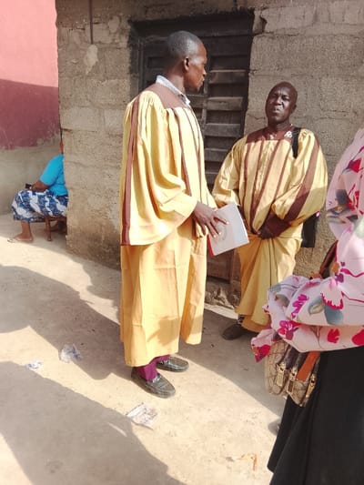 The scene depicts an outdoor gathering in Ube, Nigeria, under bright daylight conditions. Two adult males stand centrally, appearing to converse. The taller male on the left, facing right in profile, wears a long-sleeved yellow gown with brown vertical stripes and brown shoulder accents over a collared shirt. He holds a white booklet or document in his left hand. He wears black loafers and purple socks. The second male, to his right, faces forward, wearing a similar long-sleeved yellow gown with brown horizontal and vertical stripes, and a dark bag strap visible over his left shoulder. He wears dark shoes.
In the background, a rough, light grey concrete block wall is visible behind the two central males, with a dark wooden door or shuttered window recessed into it. To the left, a section of wall is painted a faded reddish-brown. A third individual, partially visible on the far left, is seated on a wooden stool, wearing a blue short-sleeved top and a blue and white patterned skirt, with light-colored sandals. A fourth individual, partially visible on the far right, wears a black skirt and a light-colored top with a pink and green floral pattern, carrying a light brown handbag with a patterned fabric and gold-colored clasps.
The ground is a light brown, possibly packed earth or concrete, showing shadows consistent with direct sunlight. Several pieces of crumpled white debris are scattered on the ground near the feet of the central figures.