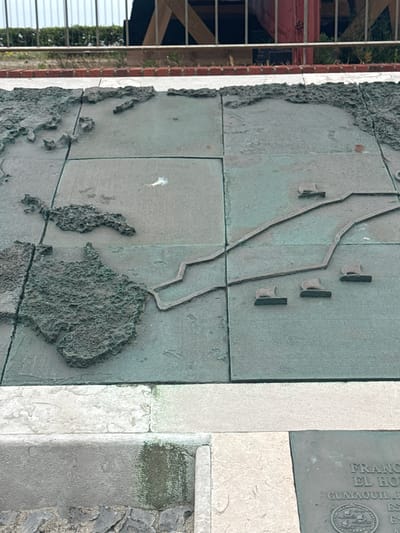 This is a close-up, slightly angled, eye-level, medium shot of a large, bas-relief bronze map mounted on a stone base. The map depicts landmasses with a textured surface and water bodies with a smooth, flat surface. Several small, raised ship figures are positioned on the water. The map appears to be divided into grid sections by etched lines. The stone base shows some staining. In the background, a metal railing and some wooden structures are visible above a brick border and green foliage. A partial view of a plaque with text, including the words "FRANC EL HO," "GUAYAQUIL," and an emblem with ships, is visible on the lower right. This is a depiction of the Monument to the Discoverers, specifically the route of the expedition of Francisco de Orellana, located in the Sardinero area of Santander, Spain.
