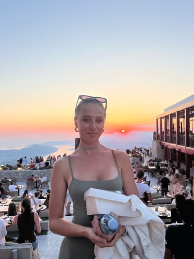 A woman in a green dress is standing in the foreground, holding a white box and a bottle of water. She is wearing sunglasses on top of her head and large hoop earrings. Behind her, a crowded outdoor dining area is set up on a terrace overlooking a bay at sunset. The sky is a gradient of orange, pink, and blue. Numerous people are seated at tables or standing, engaged in conversation. A large building with a modern design is visible on the right side of the frame. The setting appears to be a restaurant or event venue. The location is identified as Kuk, Montenegro.
