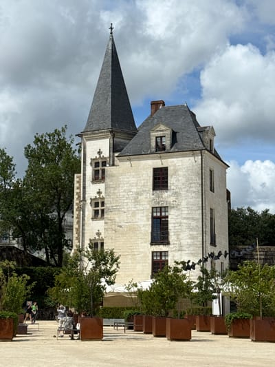 The image depicts a view of the Château des Ducs de Bretagne in Nantes, France, on a cloudy day. A tall, white stone building with a pointed, grey-shingled spire dominates the center of the frame. The building features multiple windows with dark frames, some with decorative stone tracery. To the right of the main building, a portion of a darker, pitched roof is visible, topped with a chimney.
In the foreground, a paved area with scattered benches and large, rust-colored planters filled with greenery is present. Several people are visible in the mid-ground. On the left, two individuals stand near bicycles. In the center, a group of people are seated on a bench, and another person stands nearby. Further to the right, a white canopy structure is visible behind some trees. The background is filled with lush green trees, and the sky above is a mix of grey clouds and patches of blue. A decorative string of black, bird-like figures hangs between two poles in front of the building.