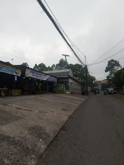 This image presents an overcast street scene in the city of Houaythat, Laos. The foreground features a dark, uneven concrete surface that transitions into an asphalt road curving slightly to the right and receding into the distance.
On the left side of the road, a series of single-story commercial buildings are visible. The most prominent structure displays two large banners; one reads "TOKO DAGING MAPAT" (Meat Shop Mapat) and the other states "SUPPLIER DAGING SAPI SEGAR & HALAL" (Fresh & Halal Beef Supplier). In front of this establishment, multiple rows of green gas cylinders are stacked on the ground. A lone individual stands near the entrance of this shop, partially obscured. Adjacent to the meat shop, another building with large glass windows showcases mannequins dressed in clothing. Further to the left, an open-fronted shop displays sacks or bags on its ground level. Overhead, a dense network of black utility lines spans across the sky, supported by a wooden utility pole that also features a solar-powered street lamp.
Beyond the buildings, lush green trees and foliage are abundant, indicating a verdant environment. On the asphalt road, three motorcycles and two cars are present. Two individuals are actively riding motorcycles away from the viewer. A black SUV or van is also visible, heading away, while a dark sedan is parked on the right side of the road further down. In the mid-distance, a banner stretched across the road displays the word "ELITE." The sky is uniformly gray and overcast, suggesting a cloudy day. The road surface exhibits signs of wear, with variations in texture and some darker, possibly wet, patches.