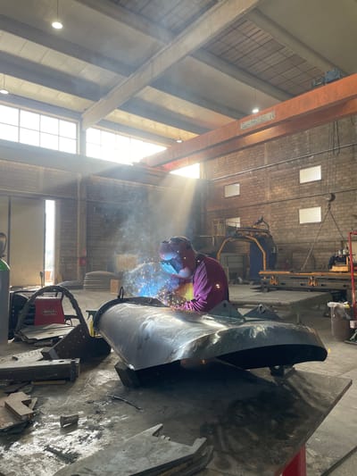 A person is welding in what appears to be a large industrial space. The individual is wearing a protective mask and red clothing and is focused on welding a large, dark metal object. Welding sparks are visible, along with smoke or fumes. The surroundings include industrial equipment and structures, such as overhead cranes and supports. Natural light streams through windows, creating bright shafts of light that diffuse through the space. The image does not provide sufficient details to confirm the location as Tomba, Montenegro.