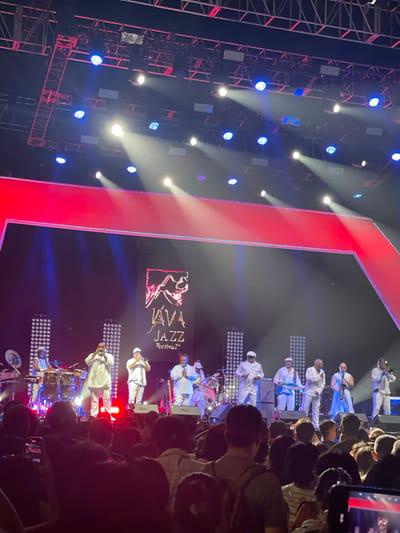 A concert is being held in Salembaran, Indonesia, with a live jazz band performing on stage in front of a crowd. The band members are dressed in white and playing various instruments, including trumpets, saxophones, guitars, and drums. The stage is lit with bright white and blue lights, and there's a large screen displaying the Java Jazz Festival logo behind the band. There is also a red vertical block on the left of the image. The crowd is dense, with people holding up their phones to record the performance. The overall atmosphere is vibrant and festive.