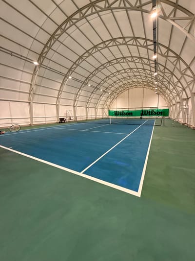 An indoor tennis court in Radomir, Bulgaria, is depicted within a large, arched structure featuring a white fabric roof supported by a gray metal truss framework. Rows of illuminated lights are mounted along the ceiling arches. The court surface consists of a blue playing area outlined by white lines, surrounded by a green perimeter. A tennis net spans the court at its midpoint. On the far side of the net, a white umpire's chair and a dark-colored bench are visible. Behind the net, a green banner bearing "Wilson" in white text twice is hung across the back wall. A dark-clad individual stands on the far side of the court, facing away from the camera, near the net. A yellow tennis ball is present on the court near the net, and another is visible in the air above the far side of the net. In the lower-left foreground, a hand holds a black tennis racket.