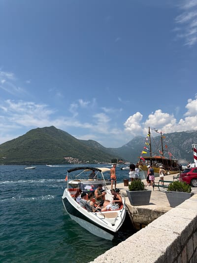 The image depicts a sunny day in the city of Perast, Montenegro, with a view of the Bay of Kotor. In the foreground, a white speed boat with an orange interior and a black canopy is docked. Several people are seated in the boat, some appearing to be tourists. To the right of the speed boat, a larger, traditional wooden boat with colorful flags is also docked. This boat appears to be a tourist vessel, with people visible on its deck.
The dock itself is made of stone, with two large planters containing green shrubs. A red car is parked to the right of the wooden boat, near a red and white striped lighthouse. In the background, the lush, green mountains of the bay rise steeply, dotted with houses. The blue water of the bay is choppy with boat wakes. The sky is a clear blue with scattered white clouds.
Several individuals are visible on the dock, some interacting with each other, and others observing the boats. A man in orange and white striped shorts stands near the speed boat. A group of people are gathered near the wooden boat, and a man in a grey shirt and pink shorts is visible. The overall scene suggests a vibrant tourist destination during a pleasant weather day.