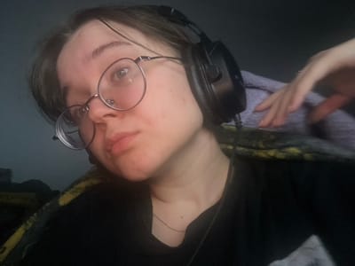 An indoor, waist-up, angled portrait features one young adult female with short, dark hair and round-frame glasses. She wears a dark t-shirt or top and a thin, light-colored necklace. Black over-ear headphones are positioned on her head, with a cable visible descending from the right earcup. Her head is tilted slightly to the viewer's left, and her gaze is directed upwards and to the viewer's right. Her expression is neutral. The subject's right hand is visible in the upper right portion of the image, resting on a purple or lavender-colored fabric. Below this, and to the subject's left, a dark fabric with a yellow or green pattern is partially visible. The background is dark and indistinct, suggesting low ambient light, with the subject's face illuminated. The specific location within Pushchino, Russia, cannot be determined from the visual information provided.