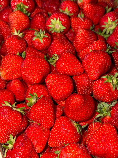 The image displays a close-up, overhead view of a large pile of ripe, red strawberries. The strawberries are a vibrant red color with visible seeds on their surface. Some of the strawberries have green stems and leaves attached. The lighting appears to be natural, casting subtle shadows on the fruit, highlighting their textured surfaces. The background is entirely filled with more strawberries, creating a dense, uniform composition. There are no people, animals, or any other objects visible in the frame. The scene is a simple and direct presentation of fresh fruit. The image does not contain any information that would allow for the identification of a specific location within the city of Aespa, Estonia, or any other geographical setting.