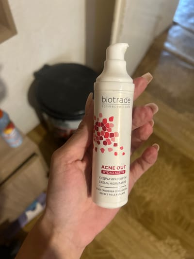 A close-up, eye-level shot shows a hand holding a white pump bottle of "ACNE OUT HYDRO ACTIVE" skincare product. The bottle features a pink and red abstract pattern near the bottom. The hand, with light skin and long fingernails, is positioned to obscure the lower portion of the bottle. In the background, slightly out of focus, is a dark, round container and a blurred object with blue and red accents. The environment appears to be indoors, with a wooden floor and a wall visible. This scene is located in Perushtitsa, Bulgaria.